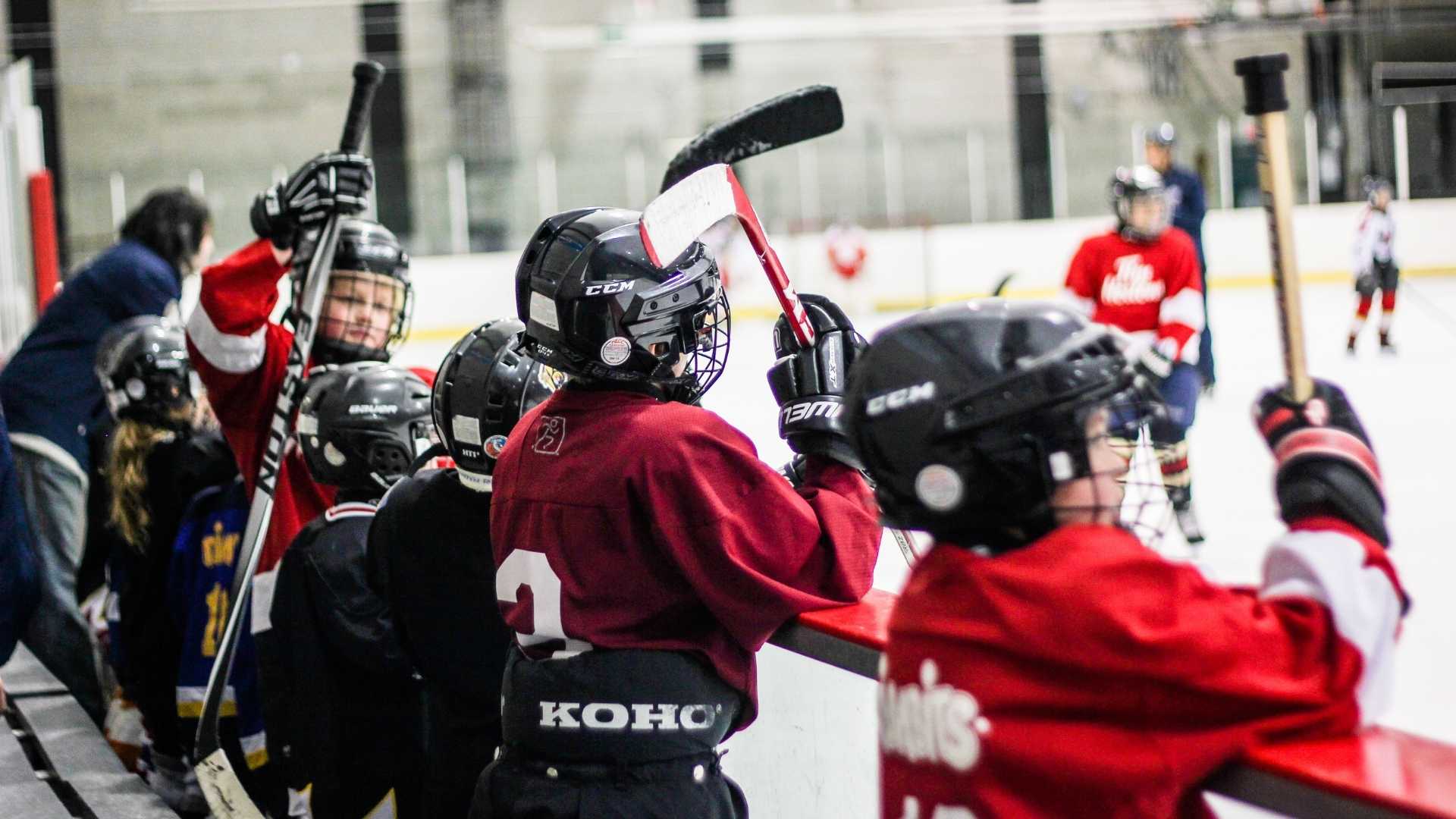 Group of kids who are wearing full hockey gears and holding their hockey sticks are standing at the side of the hockey arena while waiting for their turn to play