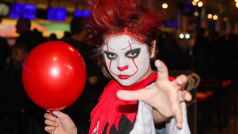 A woman dressed like a scary clown, holding a red balloon in a Halloween Bowling